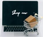 A laptop screen displaying 'Shop now' with a miniature shopping cart and cardboard box.